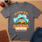 A dark gray  graphic t-shirt laid flat on a cork surface, surrounded by lifestyle props including a brown leather journal, an apple, a coffee cup, and folded blue jeans. The t-shirt features a retro-style graphic of a yellow and white van with a surfboard on its roof, set against a light blue cloud background with the text 'I AM ALREADY ON MY sUMMER VACATION. #SEEYOUINSEPTEMBER