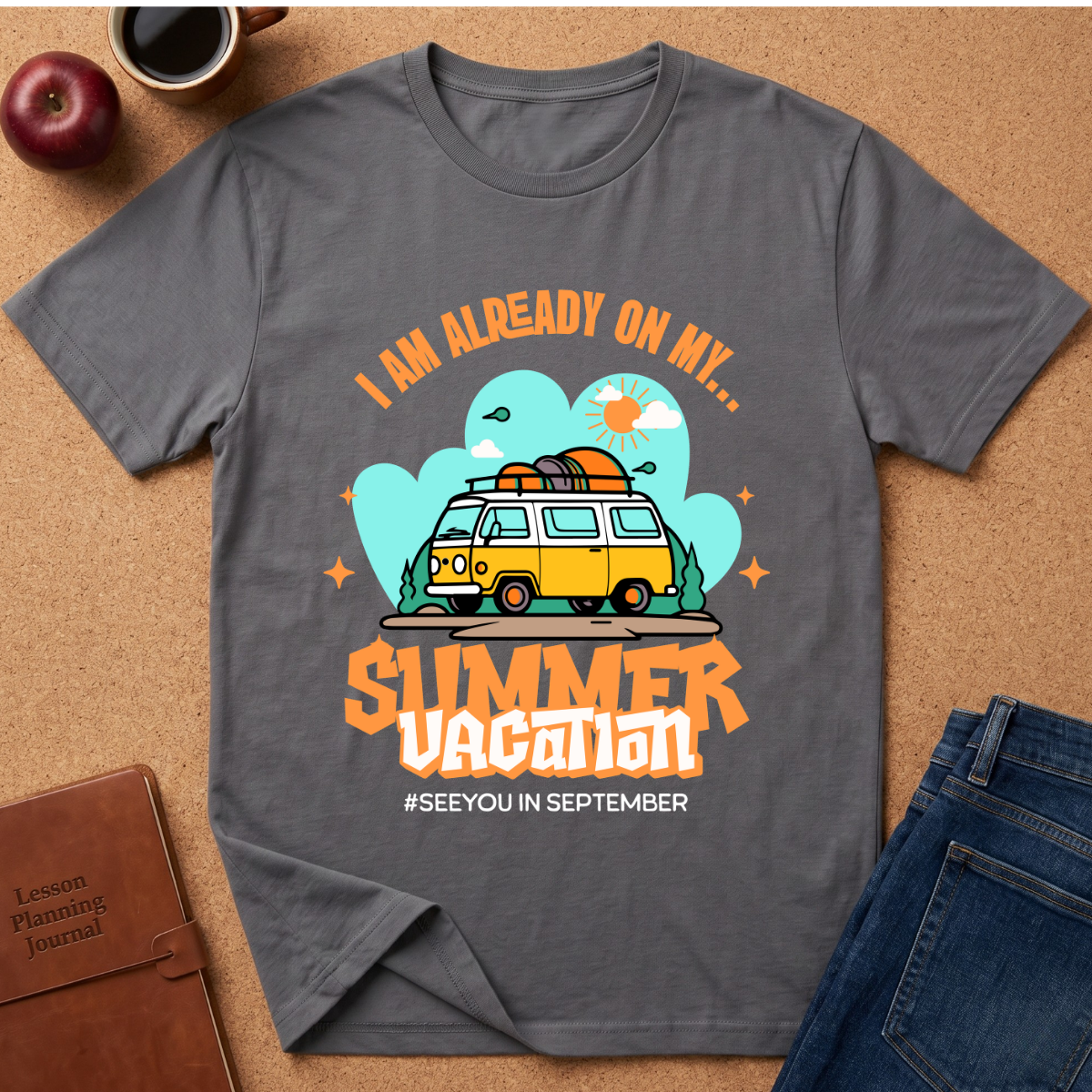 A dark gray  graphic t-shirt laid flat on a cork surface, surrounded by lifestyle props including a brown leather journal, an apple, a coffee cup, and folded blue jeans. The t-shirt features a retro-style graphic of a yellow and white van with a surfboard on its roof, set against a light blue cloud background with the text 'I AM ALREADY ON MY sUMMER VACATION. #SEEYOUINSEPTEMBER
