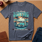 A heather slate blue graphic t-shirt laid flat on a cork background, featuring a retro surf-themed design with a VW bus, palm trees, and the text 'I'm Already SURFING'.