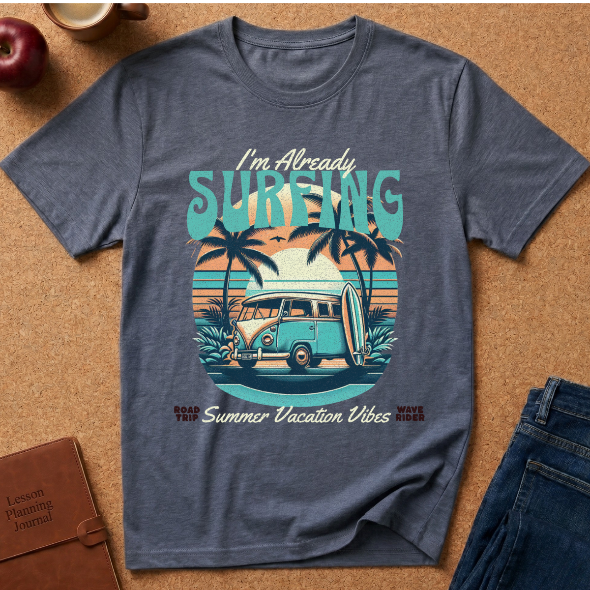 A heather slate blue graphic t-shirt laid flat on a cork background, featuring a retro surf-themed design with a VW bus, palm trees, and the text 'I'm Already SURFING'.