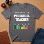 Gray t-shirt with 'Battery Life of a Preschool Teacher' graphic on a brown surface with a coffee cup, apple, and notebook.