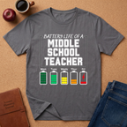 Gray t-shirt with 'Battery Life of a Middle School Teacher' graphic on a brown surface with a coffee cup, apple, and notebook.
