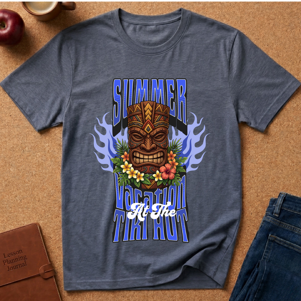A heathered slate blue t-shirt with a colorful graphic print featuring a tiki mask, flowers, flames, and the text 'SUMMER VACATION TIKI HOUR' laid flat on a cork background with lifestyle props.