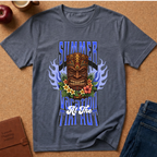 A heathered slate blue t-shirt with a colorful graphic print featuring a tiki mask, flowers, flames, and the text 'SUMMER VACATION TIKI HOUR' laid flat on a cork background with lifestyle props.