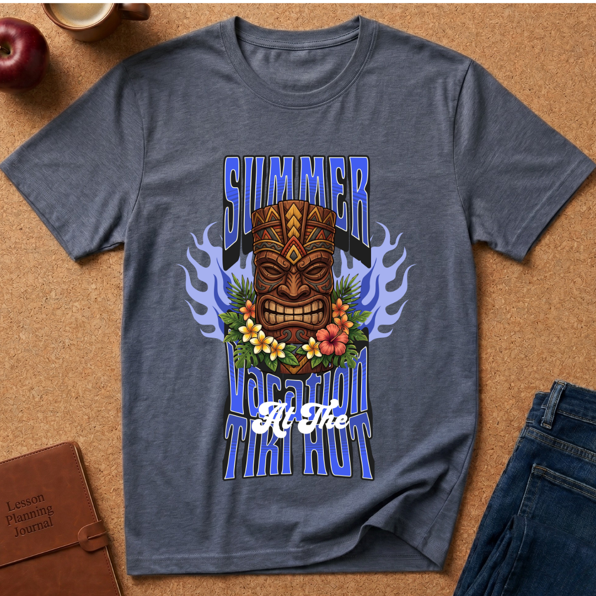 A heathered slate blue t-shirt with a colorful graphic print featuring a tiki mask, flowers, flames, and the text 'SUMMER VACATION TIKI HOUR' laid flat on a cork background with lifestyle props.