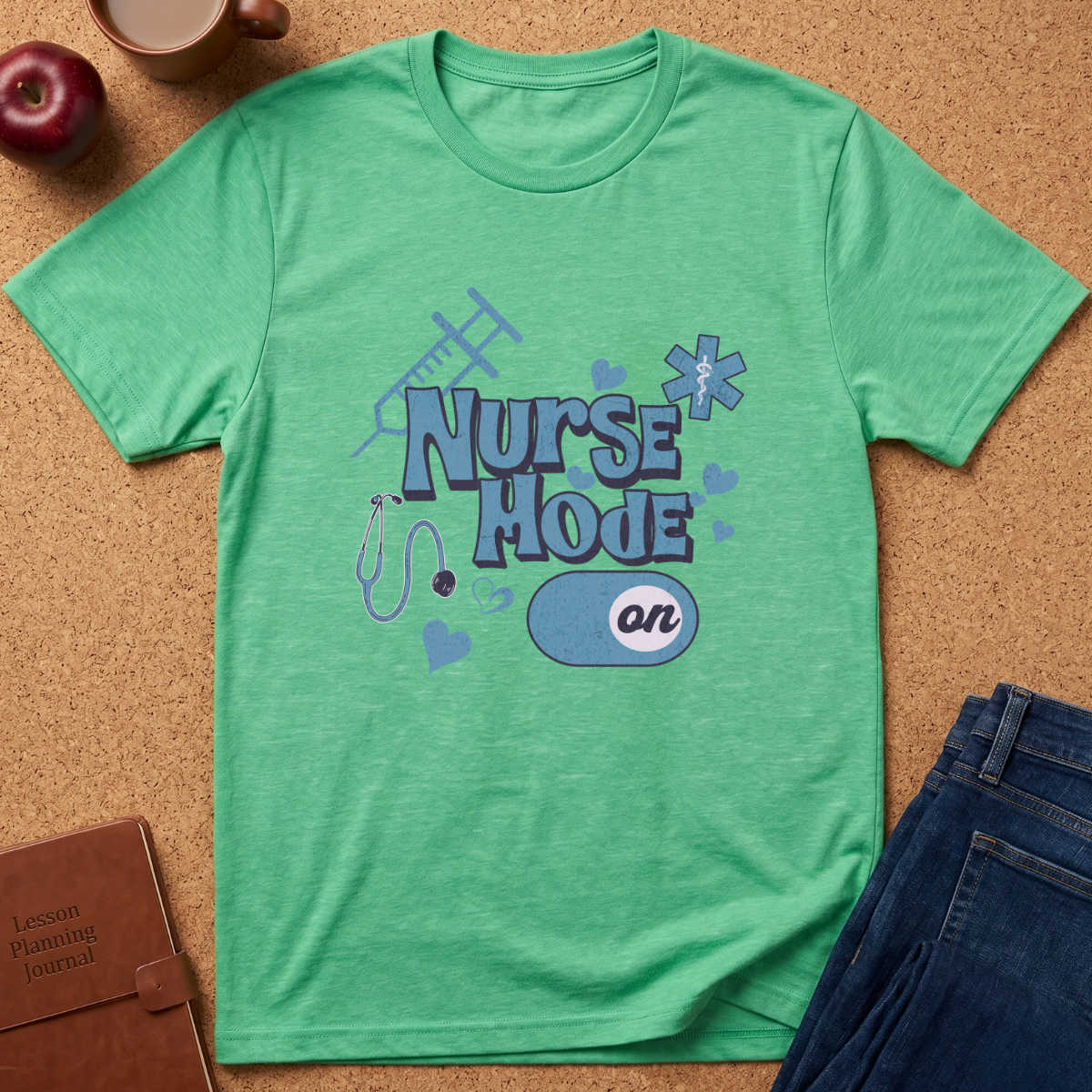 A green short-sleeve graphic T-shirt laid flat on a cork surface, featuring the words 'Nurse Mode on' with medical-themed illustrations. Styled flat-lay props include an apple, coffee cup, book, and blue jeans around the shirt.