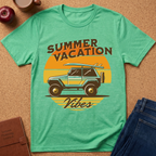 A mint green graphic t-shirt laid flat on a cork background, featuring a retro design with the text 'SUMMER VACATION' and 'Vibes' around an illustration of a blue jeep with a surfboard on its roof. Styled flat-lay props include blue jeans, an apple, a notebook, and a coffee mug.