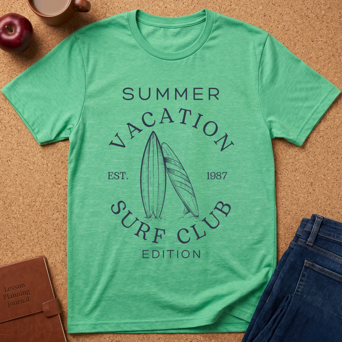 A green heather short-sleeve t-shirt with a dark navy graphic reading 'SUMMER VACATION SURF CLUB EDITION' and showing two surfboards, laid flat on a cork background with jeans, an apple, a coffee cup, and a journal nearby.