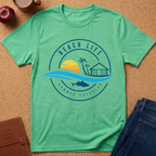 A mint green short-sleeve t-shirt with a circular 'Beach Life Summer Vacation' graphic, laid flat on a cork background with lifestyle props including an apple, a coffee mug, a notebook, and blue jeans.