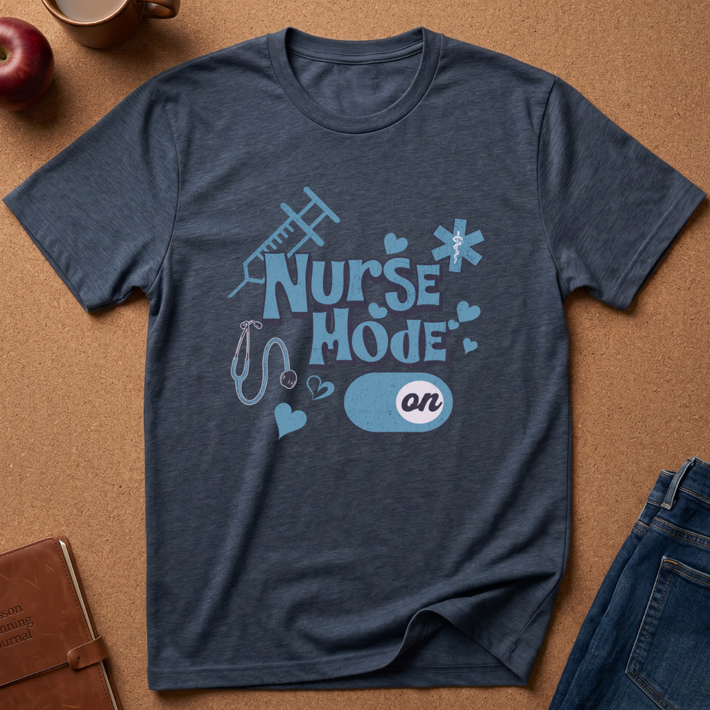 A heathered navy blue t-shirt with a 'Nurse Mode On' graphic, laid flat on a brown cork background with lifestyle props including an apple, a notebook, and jeans.