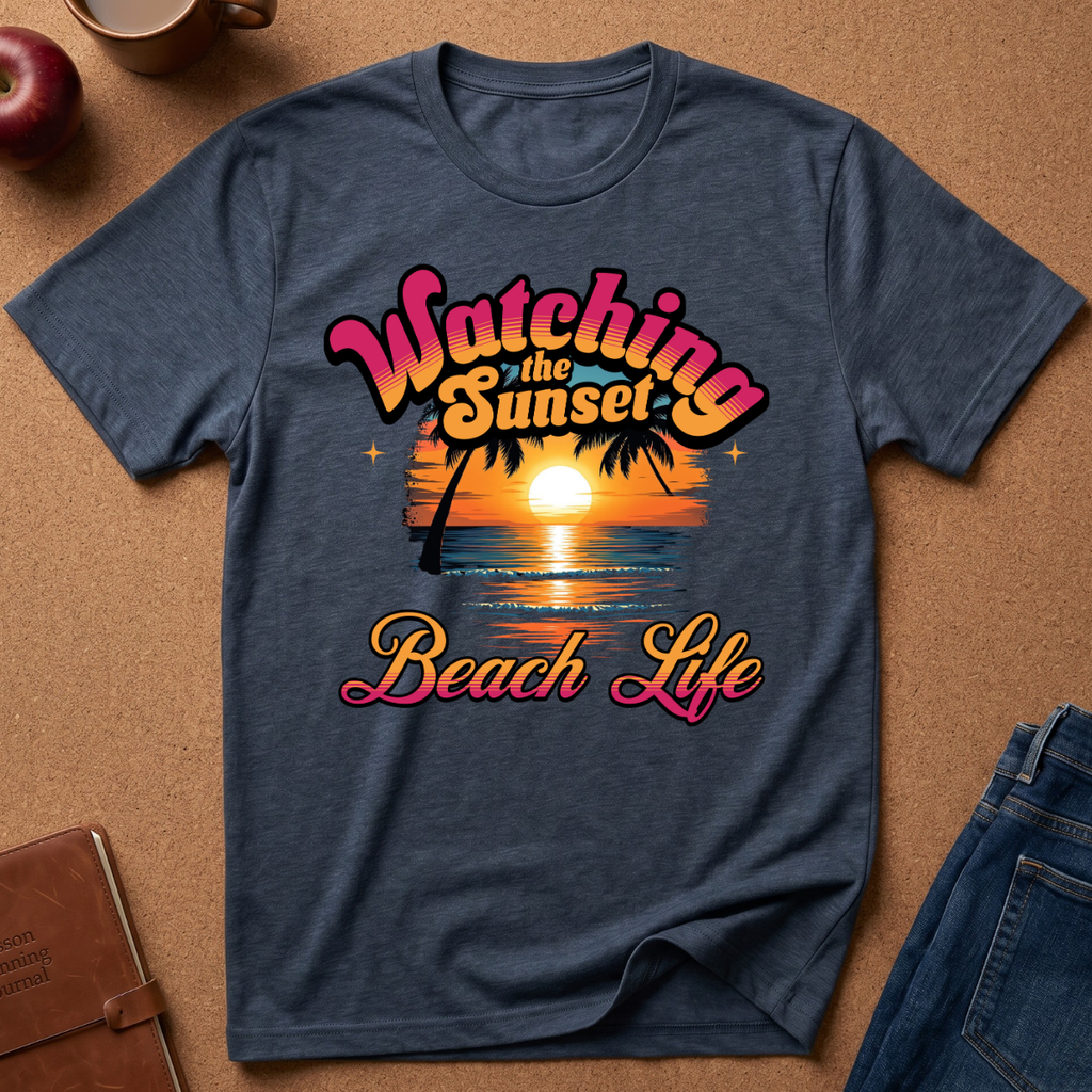 A heather navy blue t-shirt with a retro sunset graphic reading 'Watching the Sunset Beach Life' in pink, orange, and yellow, laid flat on a brown cork background with lifestyle props like jeans, a notebook, and an apple.