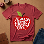 A heathered red short-sleeve t-shirt laid flat on a brown cork-like surface, featuring a large red apple graphic with the words 'TEACH INSPIRE GROW' in white and a green leaf. A coffee mug, apple, notebook, and folded jeans are placed around it as styling props.