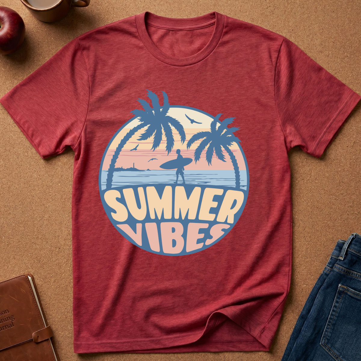 A heathered red short-sleeve t-shirt laid flat on a brown cork-like surface, featuring a circular retro graphic with palm trees, a surfer silhouette, and the words “SUMMER VIBES” in cream and blue. Partial jeans, a notebook, a cup, and an apple are visible around the shirt as styled props.