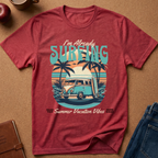 A heathered red graphic t-shirt laid flat on a brown cork background, featuring a retro-style illustration of a blue and cream van with a surfboard, palm trees, and the text 'I'm Already SURFING' and 'Dead Trip Summer Vacation Vibes Wave Rides'.