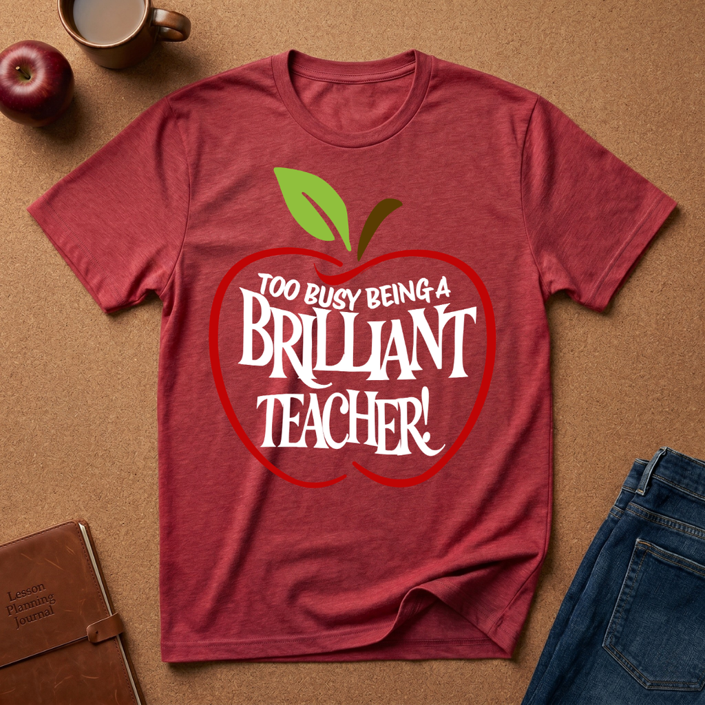 A heathered red short-sleeve t-shirt laid flat on a brown cork-like surface, featuring a large red apple graphic with green leaves and the text 'TOO BUSY BEING A BRILLIANT TEACHER!' in white. Surrounding props include a brown notebook, a coffee mug, an apple, and folded blue jeans.