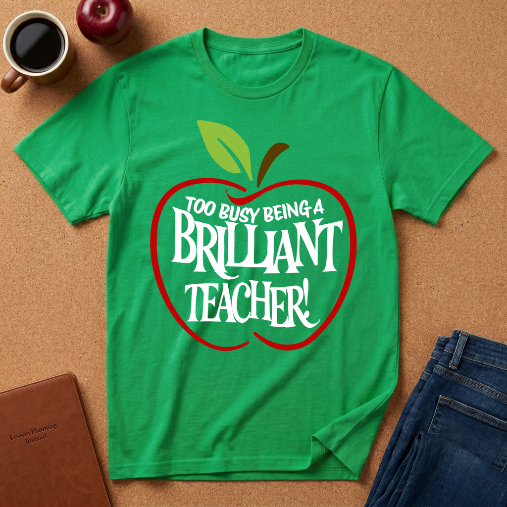 A bright green short-sleeve t-shirt with a red apple outline and the text 'TOO BUSY BEING A BRILLIANT TEACHER!' printed on the front, styled in a flat lay on a cork background with a coffee cup, apple, notebook, and jeans.