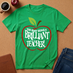 A bright green short-sleeve t-shirt with a red apple outline and the text 'TOO BUSY BEING A BRILLIANT TEACHER!' printed on the front, styled in a flat lay on a cork background with a coffee cup, apple, notebook, and jeans.