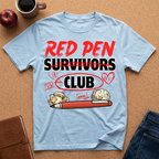 Light blue t-shirt with 'Red Pen Survivors Club' design on a brown surface with an apple, coffee, and jeans.