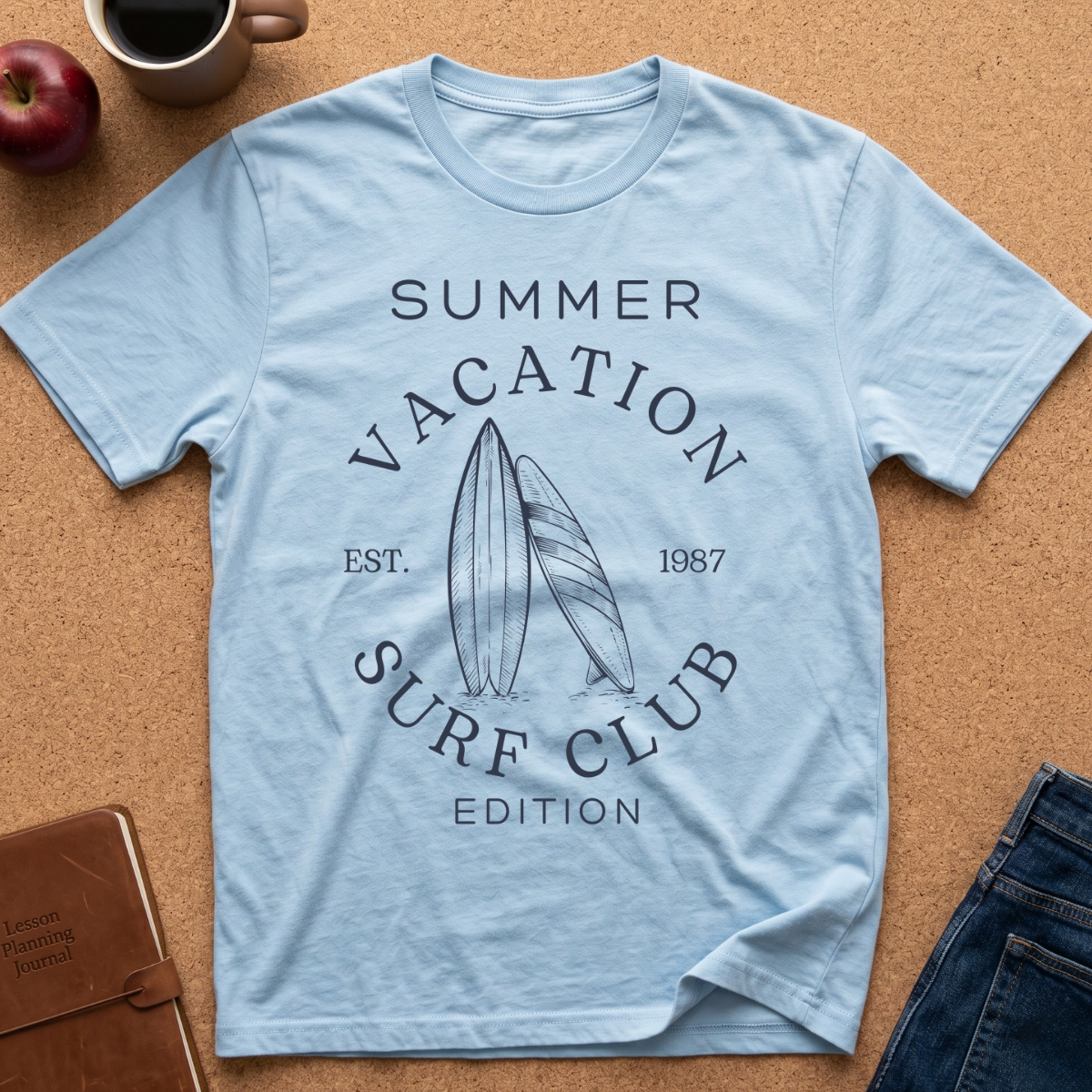 A light blue graphic t-shirt laid flat on a cork background, featuring a dark navy print that reads 'SUMMER VACATION SURF CLUB EDITION' with two surfboards and 'EST. 1987'.