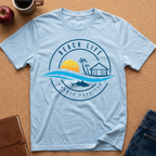 A light blue crewneck t-shirt laid flat on a cork board background, featuring a circular graphic with the text 'BEACH LIFE SUMMER VACATION' and illustrations of a sunset, palm trees, a beach hut, waves, and a shark. Styled flat-lay props include an apple, a coffee mug, a notebook, and jeans.