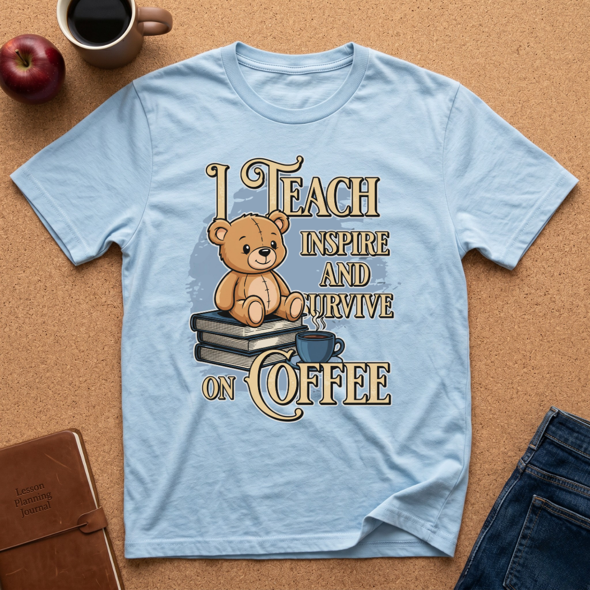 A light blue graphic t-shirt laid flat on a cork background, featuring a teddy bear sitting on books with the text 'I TEACH INSPIRE AND SURVIVE ON COFFEE'. Surrounding props include a coffee mug, an apple, a notebook, and jeans.