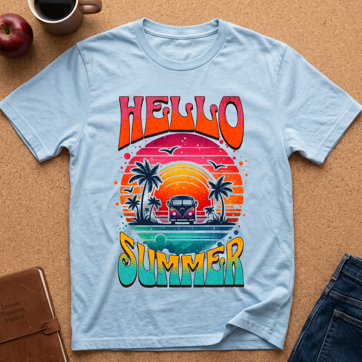 A light blue graphic t-shirt laid flat on a cork board background, featuring a retro sunset design with palm trees, a van, and the words 'HELLO SUMMER' in colorful gradient lettering. Styled flat-lay props include an apple, a coffee mug, a notebook, and jeans.
