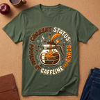 Green graphic t-shirt says "Current Status, Powered by Caffeine and Chaos" with coffee-themed graphic and text on a brown surface with apples and jeans.