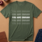 An olive green short-sleeve t-shirt with the phrase 'YOU ARE ENOUGH' printed repeatedly on the front in varying shades of gray and white. The t-shirt is laid flat on a cork background with lifestyle props including an apple, a notebook, and blue jeans.