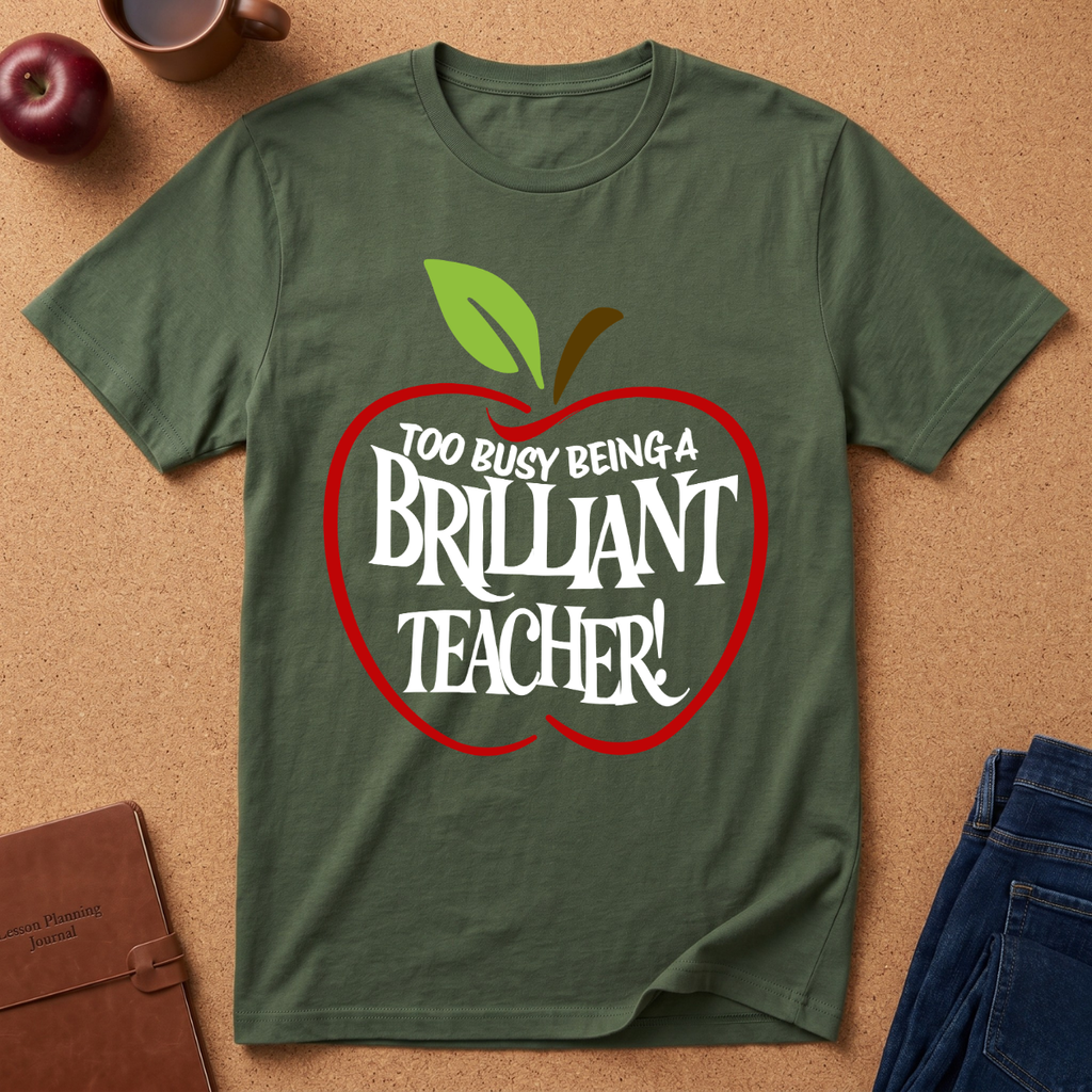 An olive green short-sleeve t-shirt laid flat on a cork background, featuring a red apple outline graphic with the text 'TOO BUSY BEING A BRILLIANT TEACHER!' in white. Styled flat-lay props include an apple, a mug, a notebook, and jeans around the shirt.