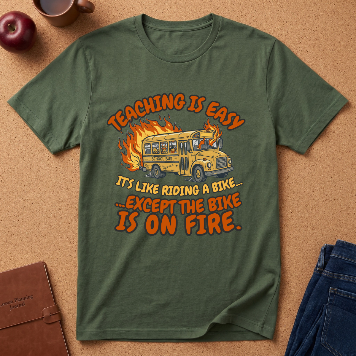 Green t-shirt with a graphic and text on a brown surface with an apple and notebook. Says Teaching is easy It's like riding a bike.. except the Bike is on fire.