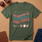 Green t-shirt with 'I'm running on attendance and vibes' text on a brown surface with an apple, notebook, and jeans.