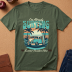 An olive green short-sleeve graphic T-shirt laid flat on a cork surface with styled props including an apple, coffee cup, notebook, and jeans. The shirt features a retro beach scene with palm trees, a van, surfboard, and the text 'I'm Already SURFING' and 'Summer Vacation Vibes'.