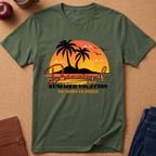 An olive green short-sleeve t-shirt laid flat on a cork background, featuring a retro sunset graphic with palm trees, birds, and the text 'Dreaming of Summer Vacation NO MORE CLASSES'.