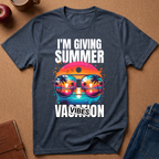 A navy blue graphic t-shirt laid flat on a brown cork background, surrounded by lifestyle props like an apple, a coffee mug, a notebook, and folded jeans. The t-shirt features a colorful graphic of sunglasses reflecting a tropical sunset with the text 'I'M GIVING SUMMER VACATION VIBES'.