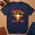 A navy blue graphic t-shirt laid flat on a cork board background, surrounded by lifestyle props including a brown leather journal, a red apple, a coffee cup, and blue jeans. The t-shirt features a retro sunset graphic with the text 'Watching the Sunset Beach Life' in pink and orange lettering.