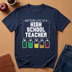 Navy blue t-shirt with 'Battery Life of a High School Teacher' graphic on a brown surface with school supplies.