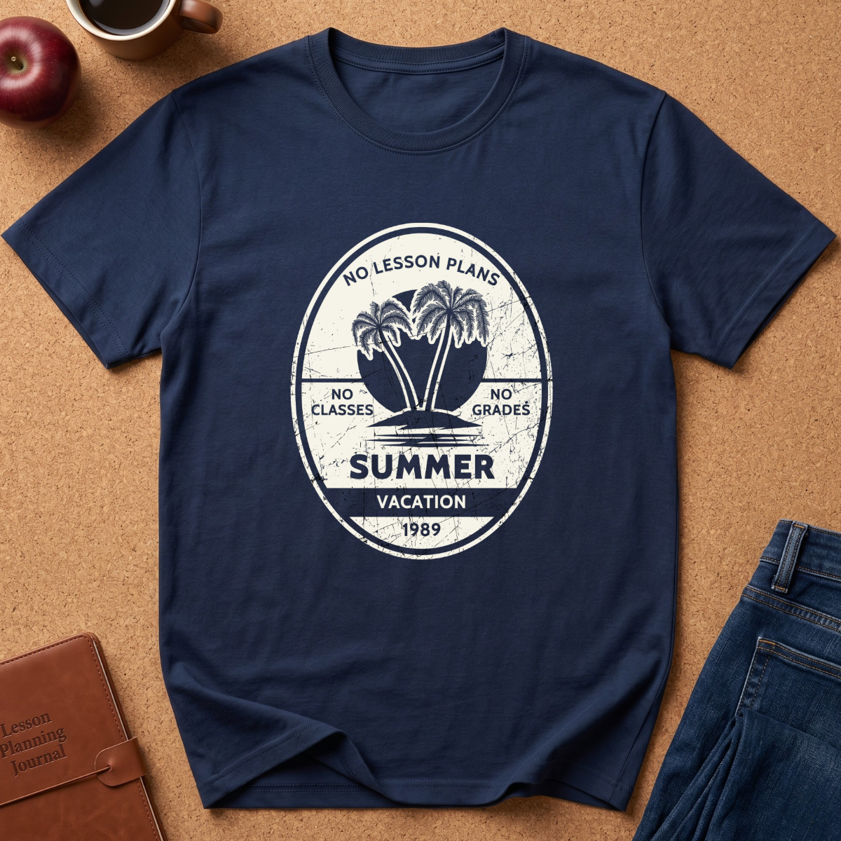 A navy blue graphic t-shirt laid flat on a cork board background, styled with a brown leather journal, an apple, and a pair of blue jeans. The t-shirt features a distressed white circular graphic with palm trees and the text 'NO LESSON PLANS', 'NO CLASSES', 'NO GRADES', 'SUMMER VACATION', and '1989'.