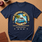 Navy Blue short-sleeve graphic T-shirt laid flat on a cork surface, featuring a retro-style beach scene with a blue van, palm tree, sun, and the text 'VAN SUMMER VACATION' and 'DRIVE INTO SUMMER WITH STYLE.' Styled props including an apple, coffee cup, notebook, and jeans are partially visible around the shirt.