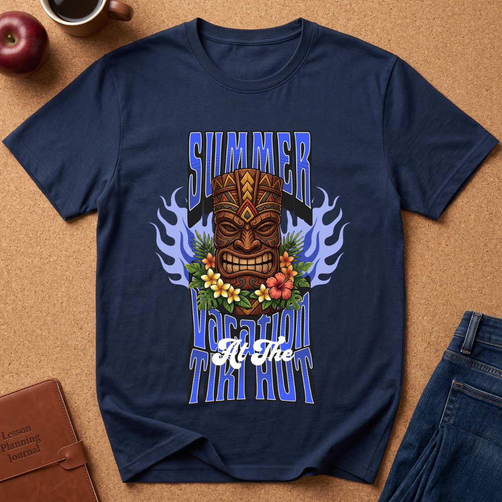 A navy blue graphic t-shirt laid flat on a cork background, featuring a tiki mask design with blue flames, flowers, and the text 'SUMMER TAI KUT At The'. Surrounding props include an apple, a coffee cup, a brown journal, and blue jeans.