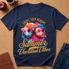 A navy blue short-sleeve t-shirt laid flat on a cork background, featuring a colorful graphic of sunglasses with palm tree reflections and the text 'Enjoy Your Moment Summer Vacation Vibes' in yellow and orange. Styled flat-lay props include a brown leather journal, a blue denim jacket, a red apple, and a coffee cup.