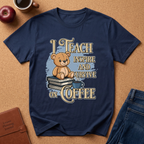 A navy blue graphic t-shirt laid flat on a cork background, featuring a cartoon teddy bear sitting on books with the text 'I TEACH INSPIRE AND SURVIVE ON COFFEE'. Styled flat-lay props include an apple, a coffee cup, a leather journal, and blue jeans.