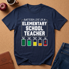 Navy blue t-shirt with 'Battery Life of a Elementary School Teacher' graphic on a brown surface with school supplies.
