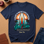 A navy blue graphic t-shirt laid flat on a brown cork background, surrounded by lifestyle props including a coffee cup, an apple, a notebook, and jeans. The t-shirt features a retro surfboard design with the text 'HAVE A NICE VACATION Good Day' and a humorous vacation-themed quote.
