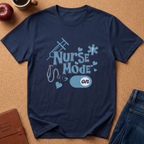 A navy blue short-sleeve t-shirt with a light blue graphic reading 'Nurse Mode on' surrounded by medical-themed icons, laid flat on a brown cork background with props including an apple, a coffee mug, a journal, and jeans.