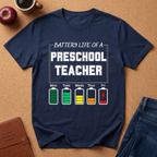 Navy blue t-shirt with 'Battery Life of a Preschool Teacher' graphic on a brown surface with school supplies.