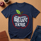 A navy blue graphic t-shirt laid flat on a cork background, featuring a red apple outline with the text 'TOO BUSY BEING A BRILLIANT TEACHER!' in white. The shirt is styled with a brown leather journal, a coffee cup, an apple, and blue jeans.