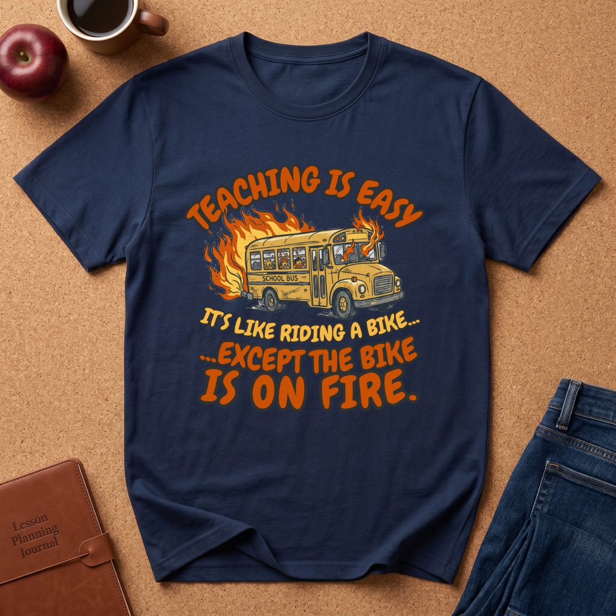 Navy blue t-shirt with a graphic of a school bus on fire and humorous text, placed on a brown surface with an apple and notebook. Teaching is easy It's like riding a bike.. except the Bike is on fire.