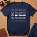 A navy blue graphic t-shirt laid flat on a cork background, featuring the phrase 'YOU ARE ENOUGH' printed in stacked rows, with the middle line in solid white and the others in faded gray. Styled flat-lay props include an apple, a coffee cup, a journal, and jeans.