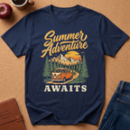 A navy blue graphic t-shirt laid flat on a brown cork background, featuring a retro-style 'Summer Adventure AWAITS' design with a vintage orange bus, mountains, pine trees, and a yellow sun. Styled flat-lay props include an apple, coffee cup, leather journal, and blue jeans.
