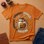 Orange graphic t-shirt says "Current Status, Powered by Caffeine and Chaos" with coffee-themed graphic and text on a brown surface with a coffee cup and apple.